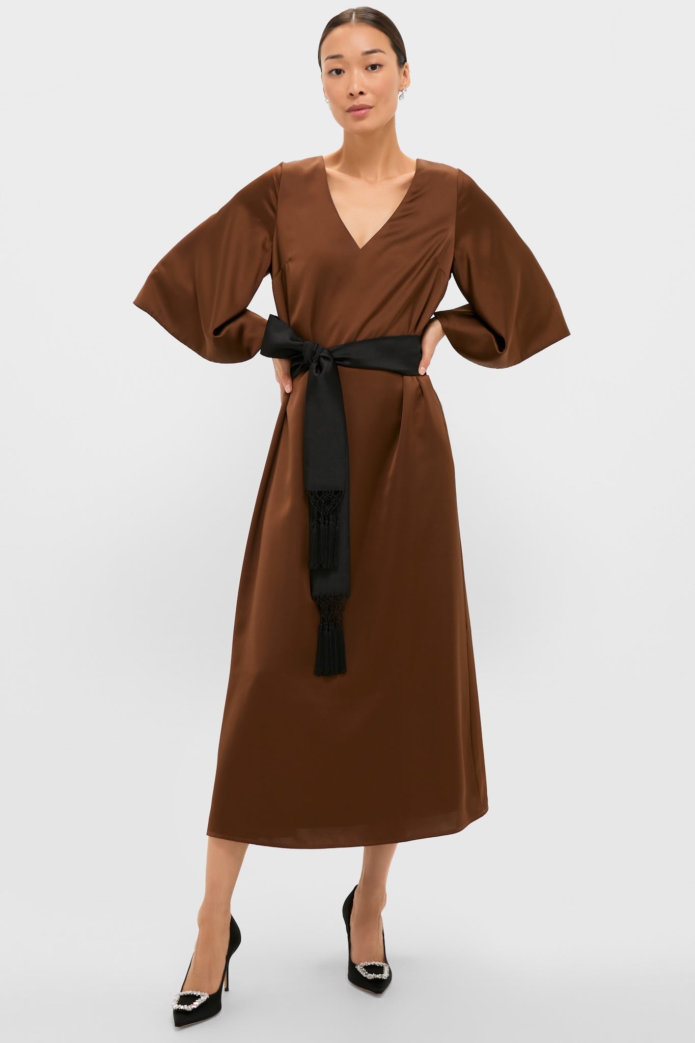 Chocolate Brown Satin Emiliana Maxi Dress with a classic V-neck and wide bell sleeves, featuring an effortless black tassel-tied belt. Paired with high-end pointed-toe heels.
