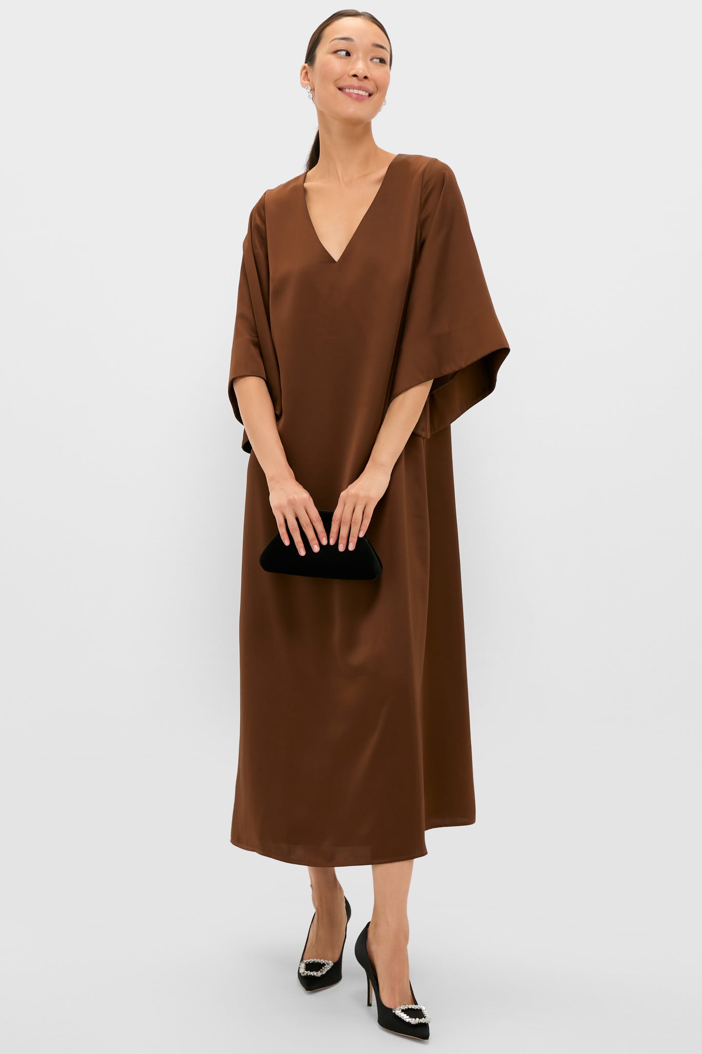 Woman in a Chocolate Brown Satin Emiliana Maxi Dress with kimono sleeves, accessorized with crystal-buckle stiletto pumps and a clutch, exuding classic, effortless high-end style.