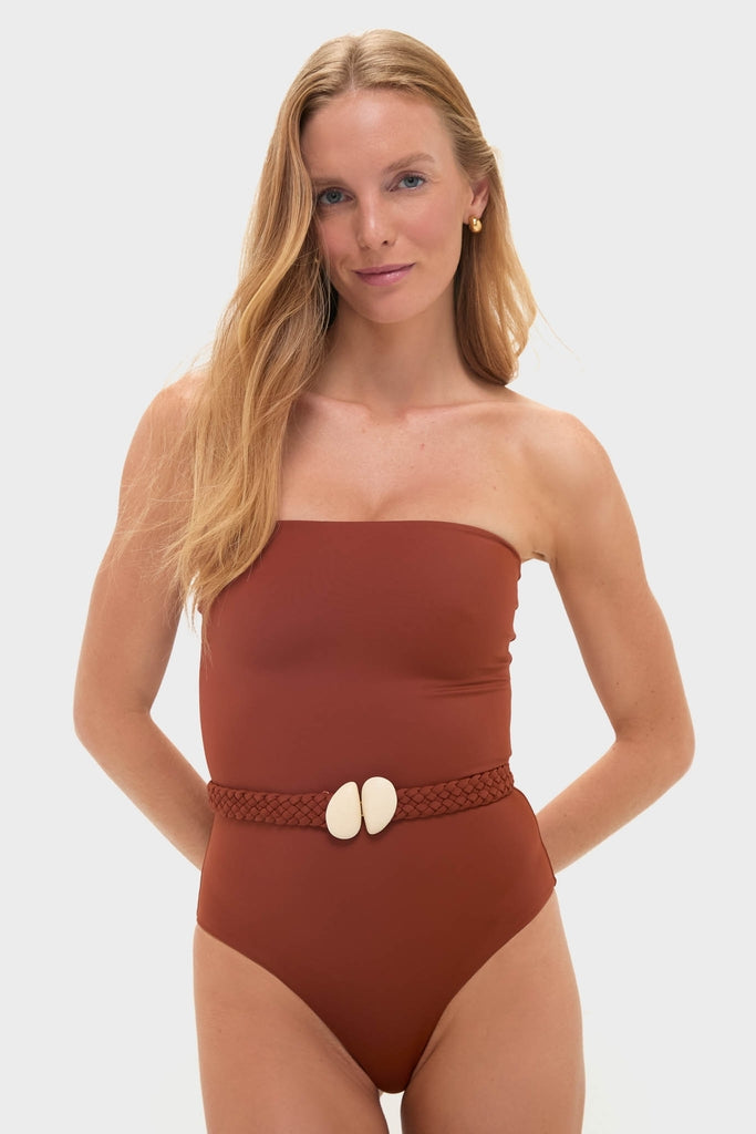 Woman models Marsala The Classic One Piece swimsuit in rust-brown, featuring a strapless silhouette, removable braided belt, and shell details; effortlessly chic and fun.