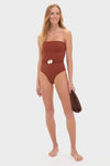 Woman in rust-brown strapless swimsuit with removable braided belt and shell details, holding a dark brown clutch, embodying classic, effortless, high-end style.
