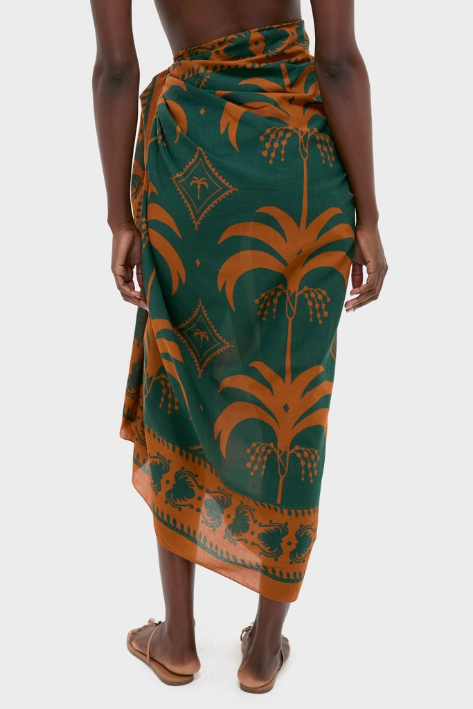 Rust and Dark Green Dance of Pareo, a classic asymmetric wrap skirt with a palm-tree motif, paired with effortless flat sandals for a high-end, fun look.