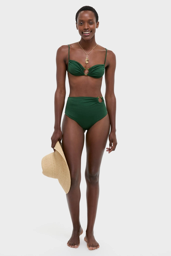Model in a dark green Sumatra Green Taita Bikini Bottom with ring detail, holding a straw sun hat, exuding an effortless, high-end, and fun vibe.