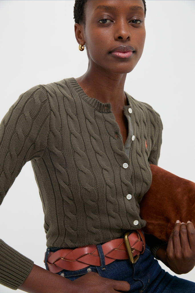 Classic cable-knit Harvest Olive Cardigan, buttoned, showcasing effortless style with ribbed trim and signature pony, paired with jeans and brown accessories for a fun, high-end look.