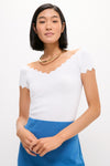 Woman wearing a classic, effortless White Off-the-Shoulder Ric Rac Compression Elliston Shirt with scalloped trim, paired with a chunky gold necklace and high-waisted blue bottom.