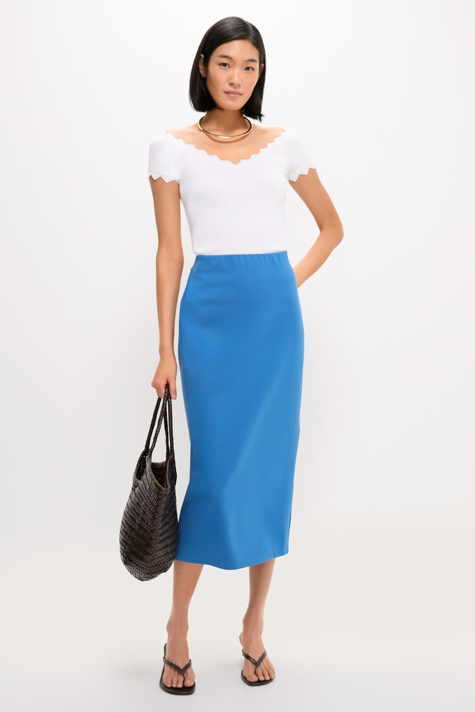 Model wearing the classic White Off-the-Shoulder Ric Rac Compression Elliston Shirt, styled effortlessly with a blue midi skirt and gold necklace, embodies high-end, fun fashion.