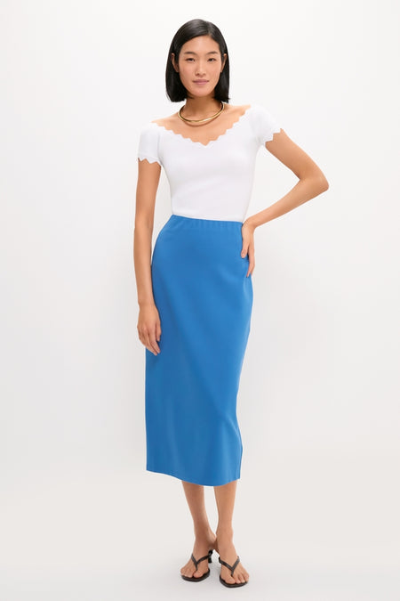 Young woman in a classic, effortless White Off-the-Shoulder Ric Rac Compression Elliston Shirt, paired with a vibrant blue skirt and gold necklace, embodying high-end, fun fashion.