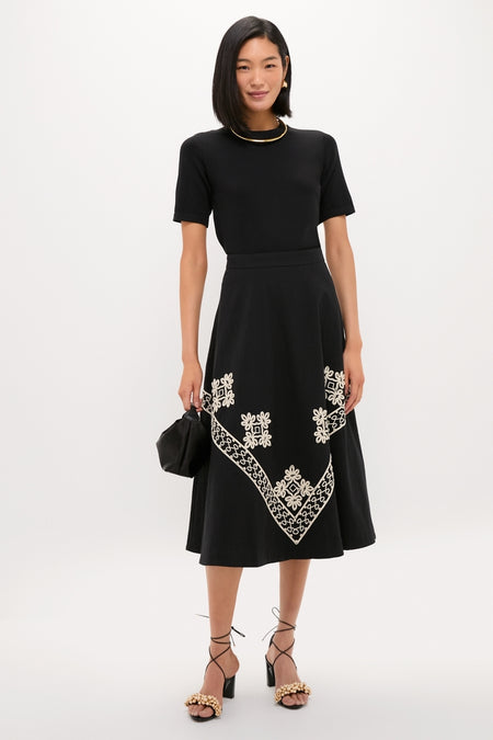 Model showcasing the classic Soutache Cotton Andrea Circle Skirt, featuring an effortless black midi design with a white floral chevron border, paired with a chic pouch handbag.