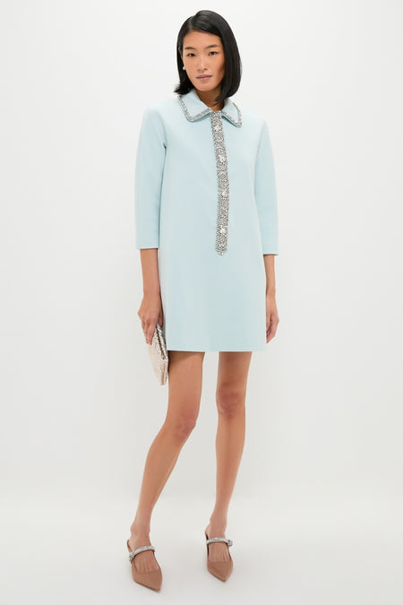 Woman in Ice Blue Embellished Nicola Dress, showcasing a classic, effortless style with a jeweled collar and placket, paired with a metallic clutch and rhinestone mules.