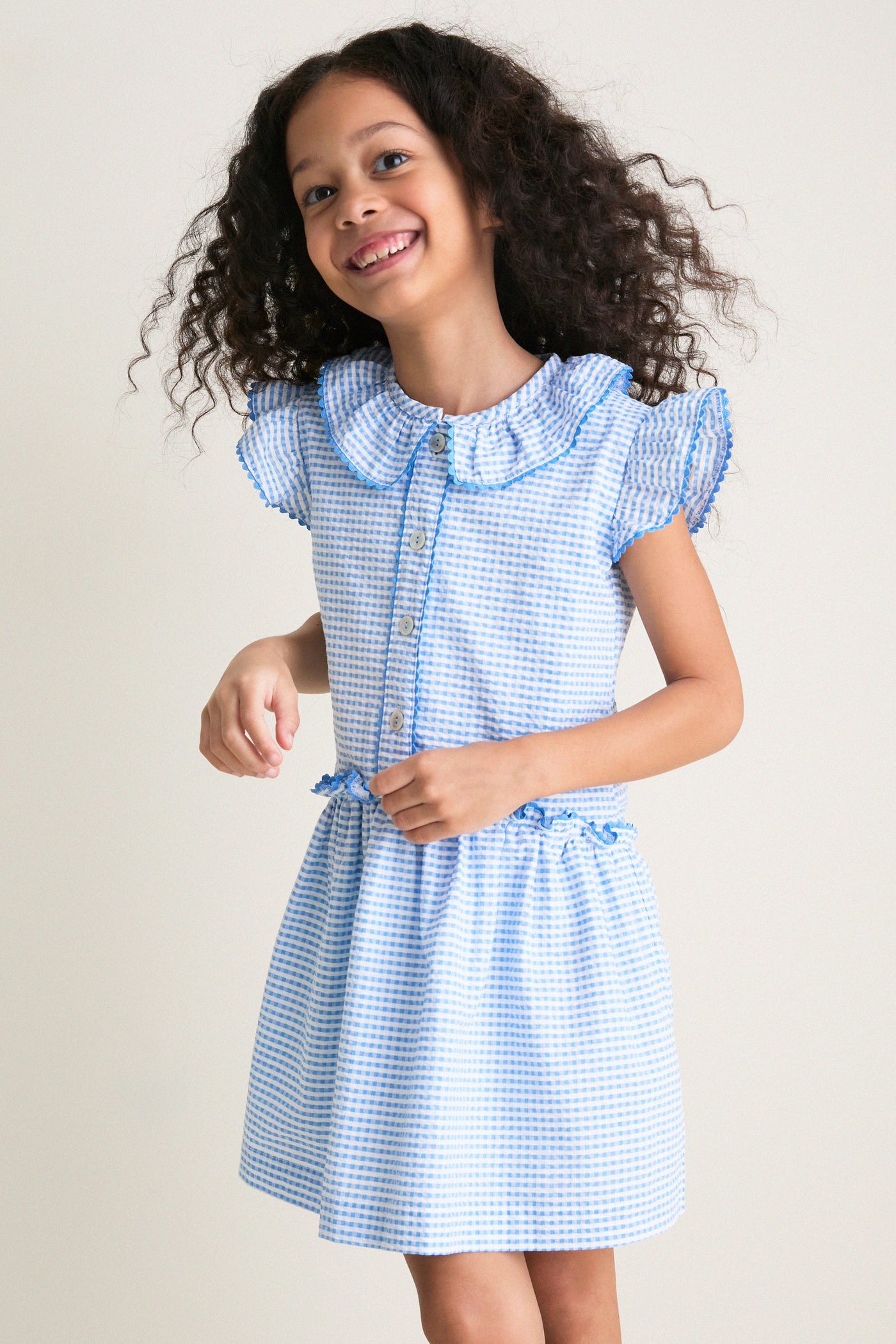 Young girl wearing the classic Blue Gingham Fabiola Drop Waist Dress, featuring an effortless, fun design with butterfly sleeves, a frill collar, and high-end detailing.