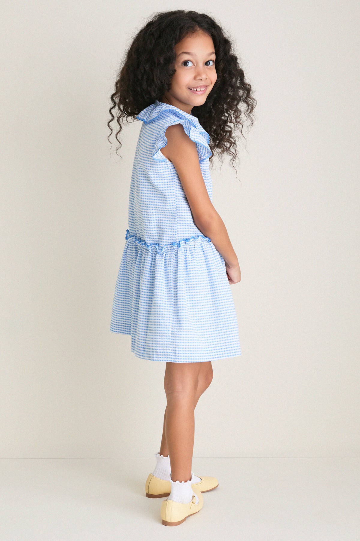 Young girl in profile wearing the Blue Gingham Fabiola Drop Waist Dress with butterfly sleeves, embodying classic, effortless style. Perfect for fun, everyday moments and high-end occasions.