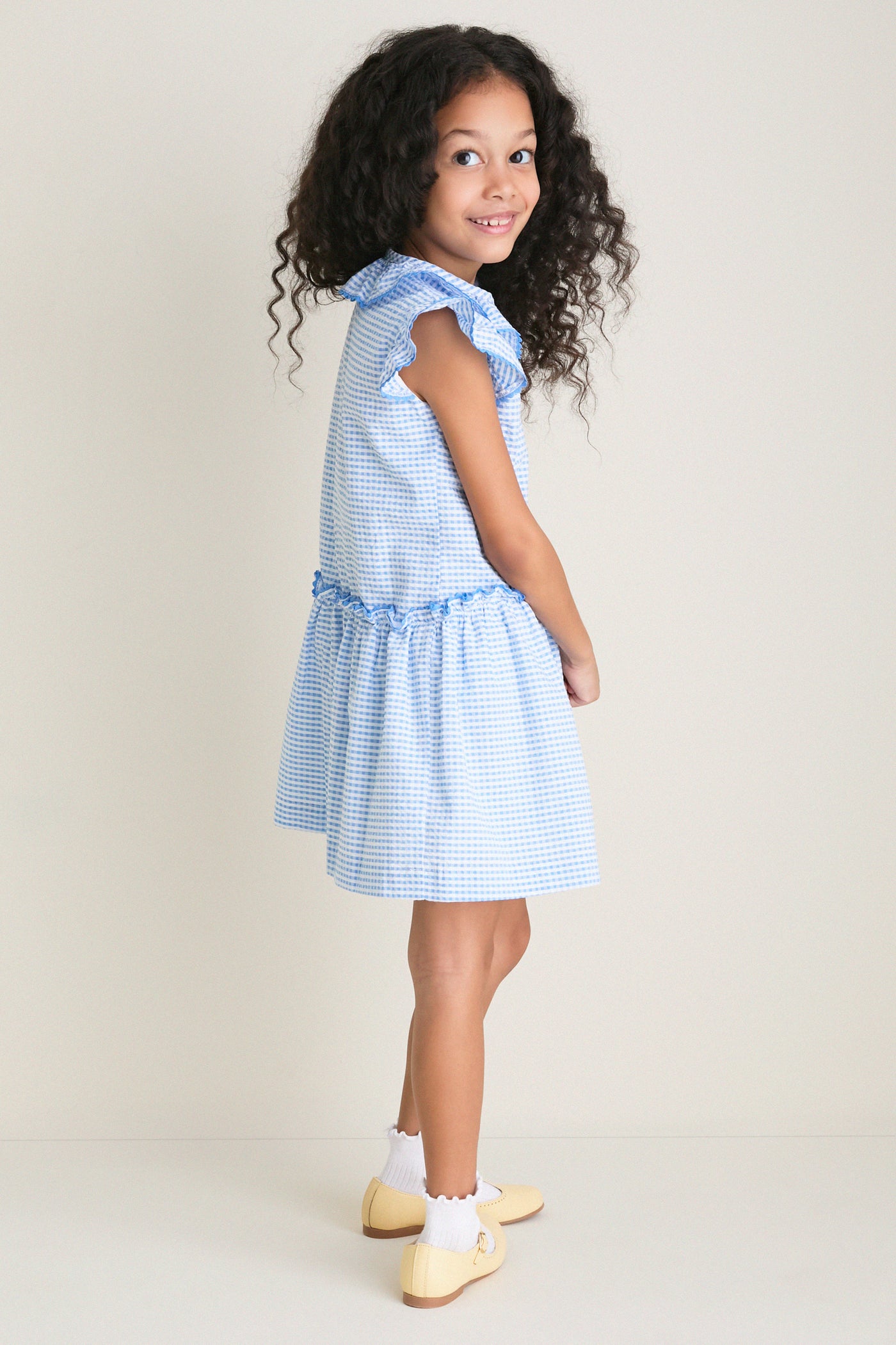 Young girl in profile wearing the Blue Gingham Fabiola Drop Waist Dress with butterfly sleeves, embodying classic, effortless style. Perfect for fun, everyday moments and high-end occasions.