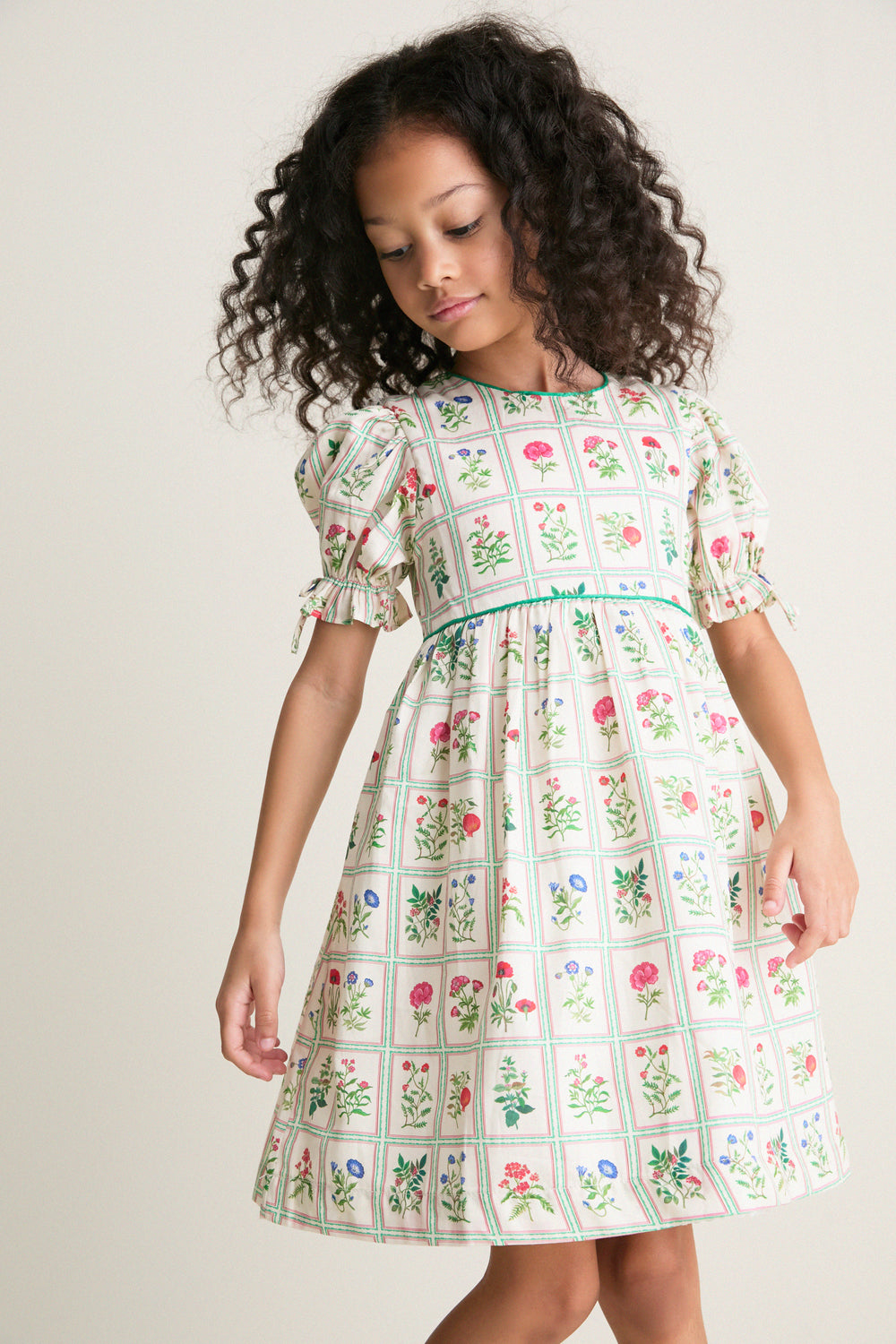 Young girl in a classic, high-end Ivory Botanical Olivia Dress, showcasing effortless charm with puffed sleeves, floral grid print, and fun, feminine details.