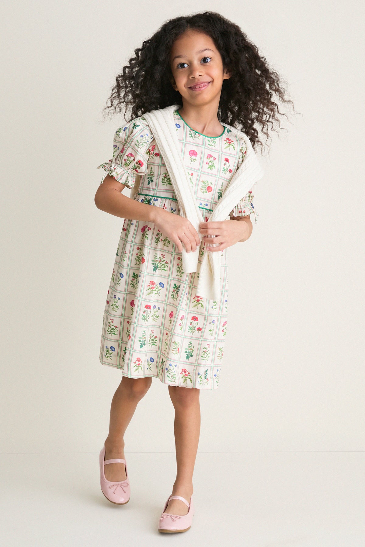Young girl in an Ivory Botanical Olivia Dress, featuring classic floral prints and a high-end empire waist, effortlessly styled with a draped sweater and fun Mary Jane shoes.