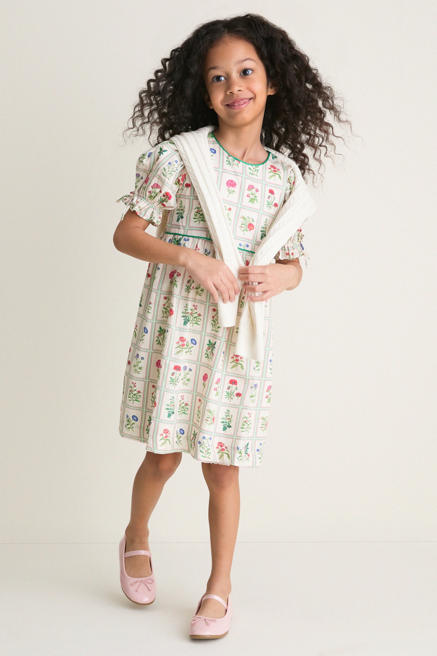 Young girl in an Ivory Botanical Olivia Dress, featuring classic floral prints and a high-end empire waist, effortlessly styled with a draped sweater and fun Mary Jane shoes.