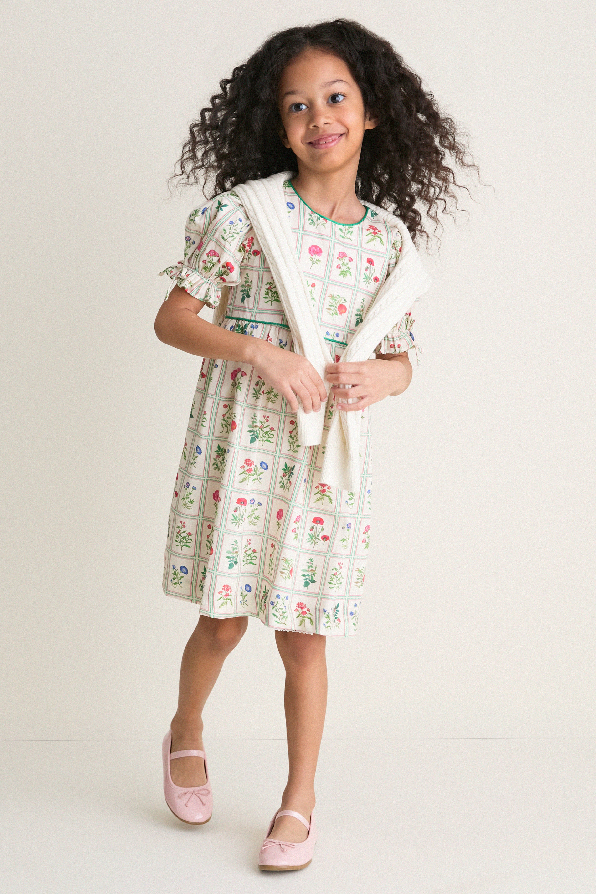 Young girl in an Ivory Botanical Olivia Dress, featuring classic floral prints and a high-end empire waist, effortlessly styled with a draped sweater and fun Mary Jane shoes.