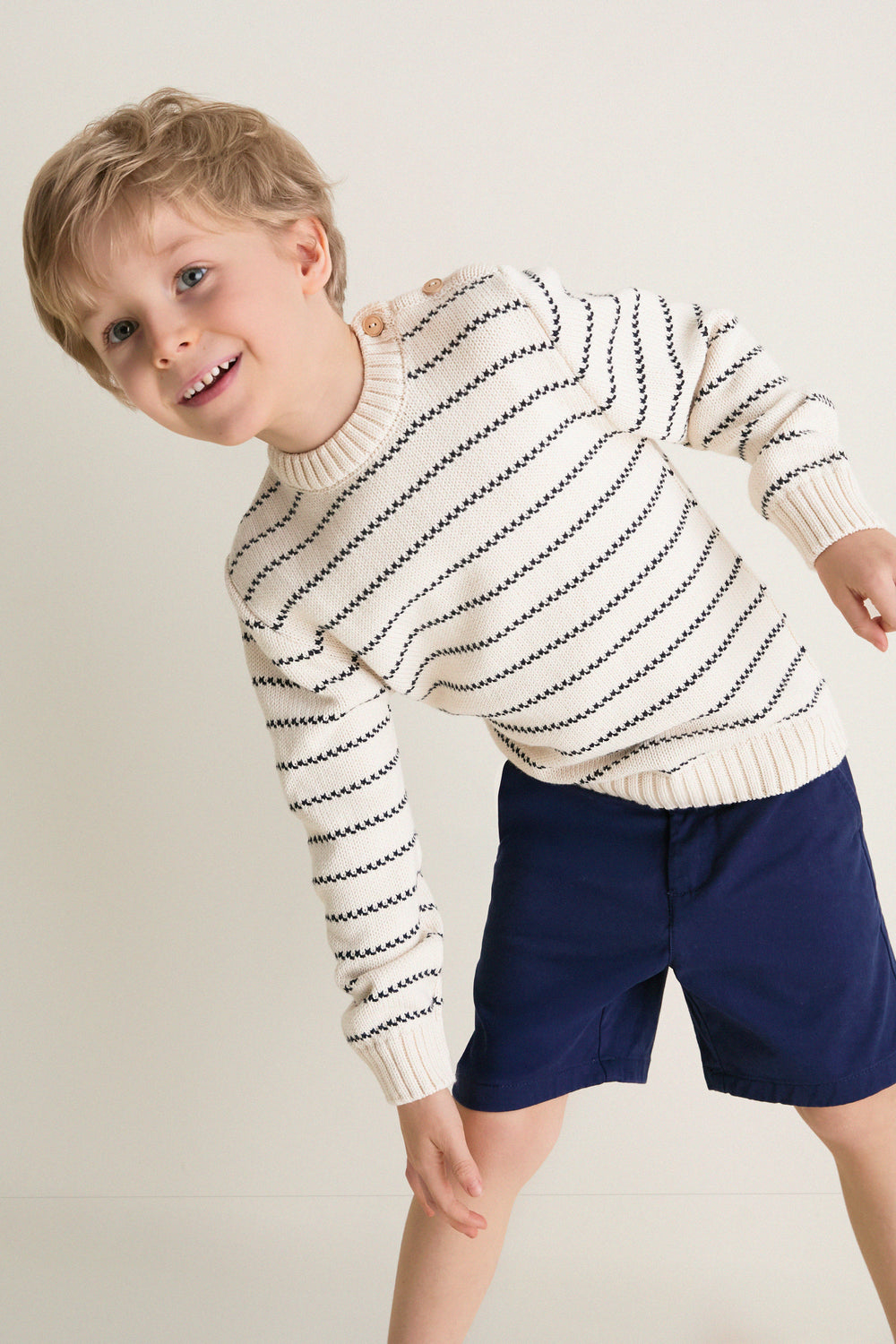 Young boy smiling in a classic Navy Stripe Raya Cotton Jumper, featuring ribbed trims and wooden shoulder buttons. Effortless and fun, perfect for high-end style.