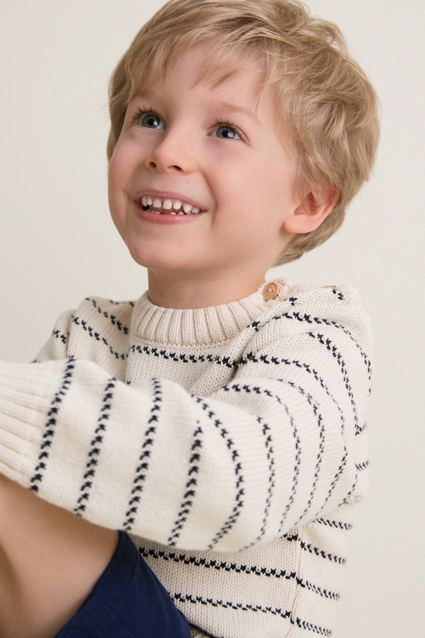 Young boy in a Navy Stripe Raya Cotton Jumper, smiling brightly. The classic, effortless design features wooden shoulder buttons, embodying high-end, fun style.