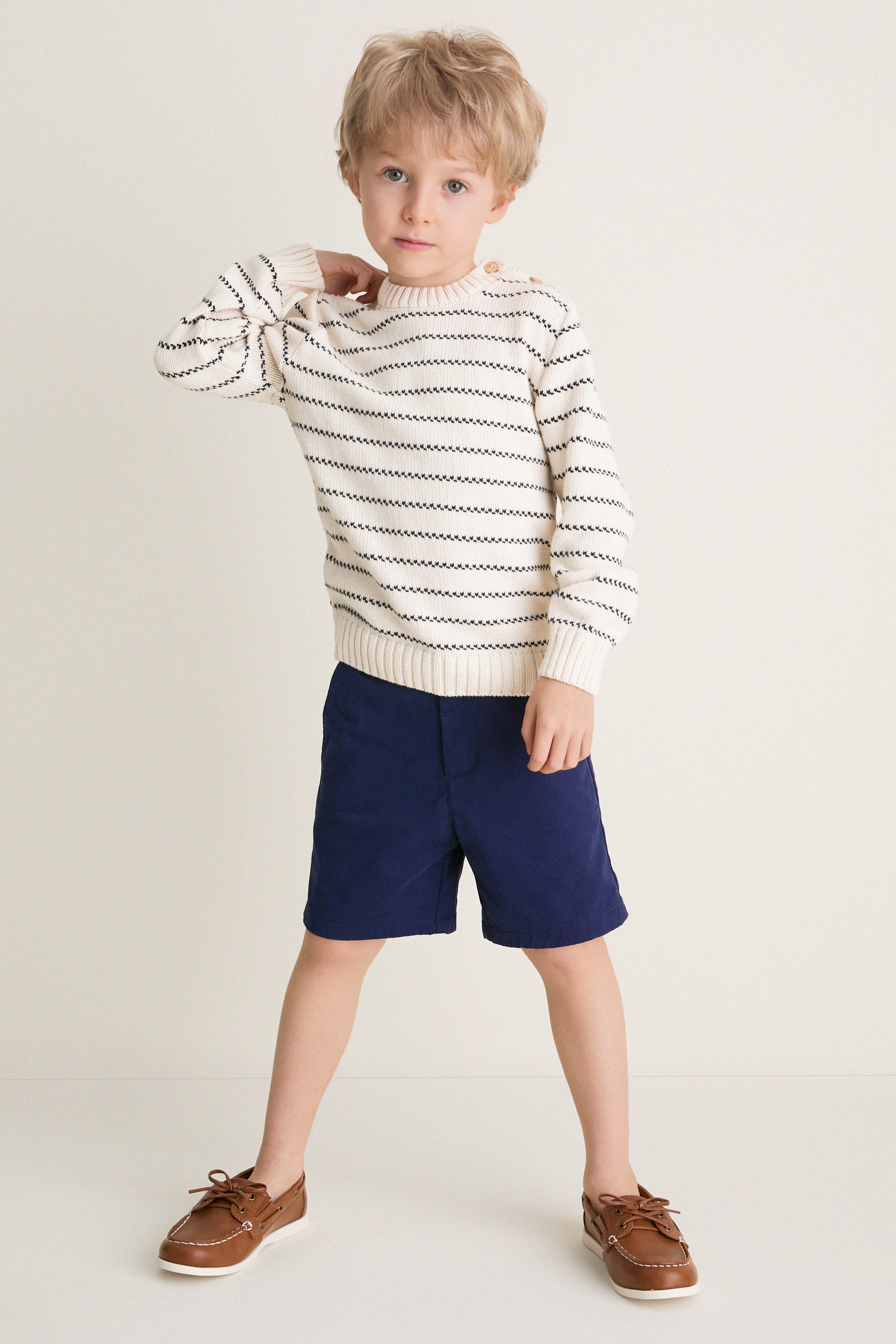 Young boy in a classic Navy Stripe Raya Cotton Jumper, showcasing effortless style with ribbed trims and wooden buttons, paired with navy shorts and brown boat shoes.