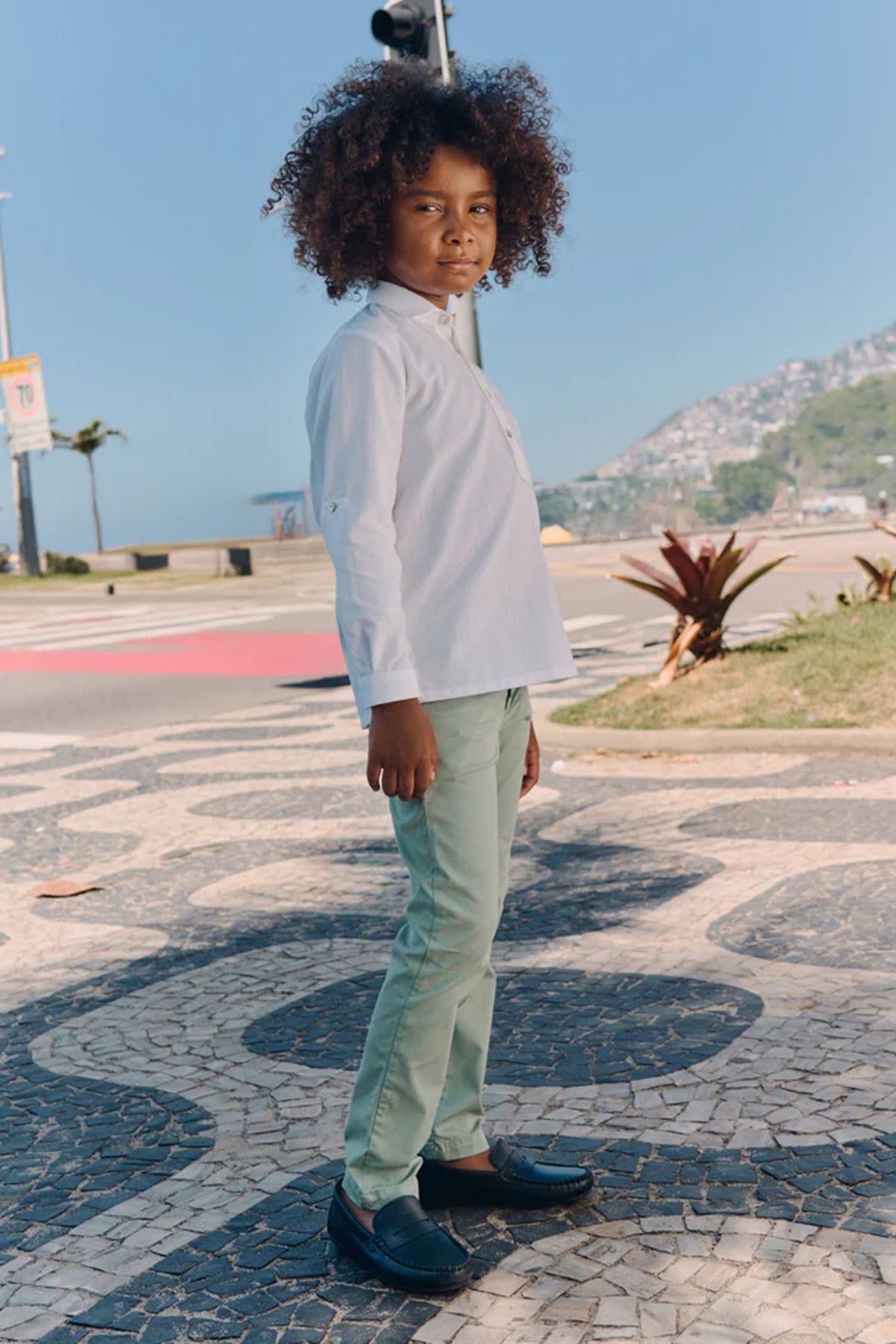 Young boy wearing classic Sage Green Elio Chinos with a white shirt, standing on a mosaic pavement, embodying effortless and high-end style, perfect for fun, everyday adventures.