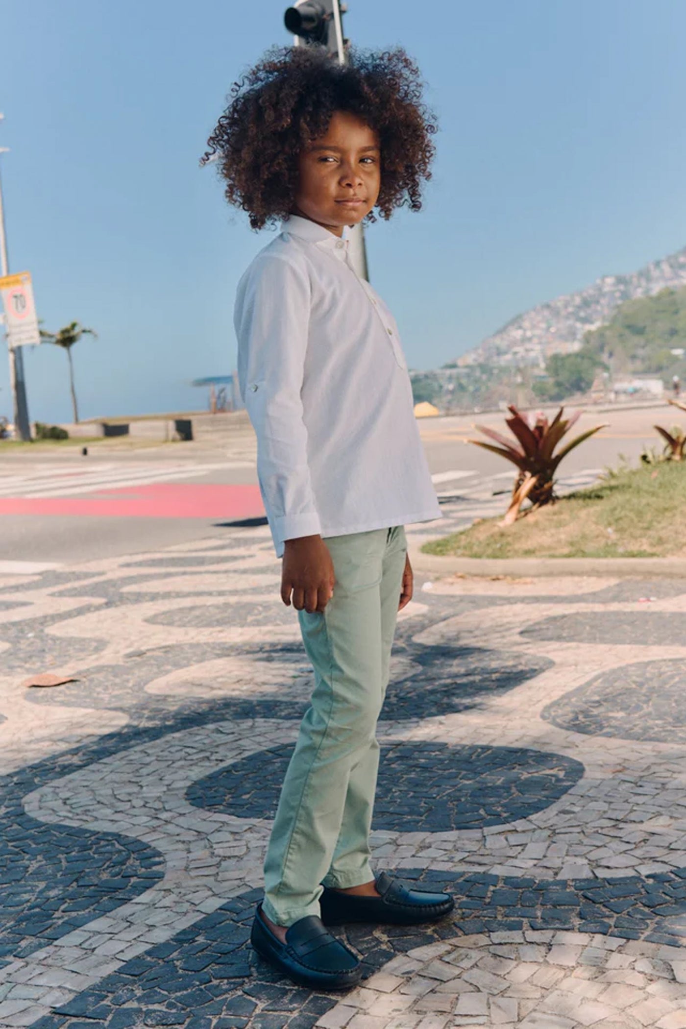 Young boy wearing classic Sage Green Elio Chinos with a white shirt, standing on a mosaic pavement, embodying effortless and high-end style, perfect for fun, everyday adventures.