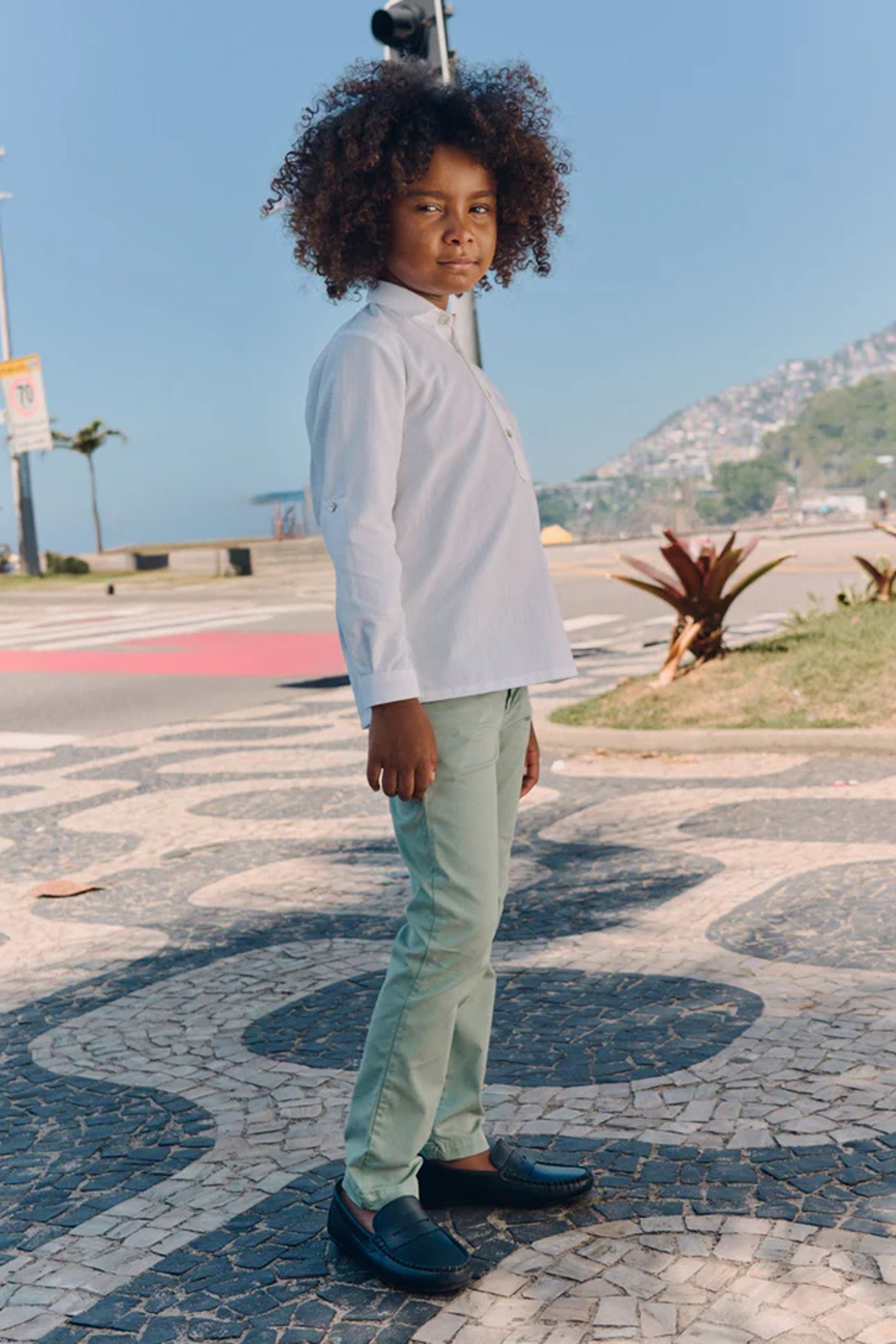Young boy wearing classic Sage Green Elio Chinos with a white shirt, standing on a mosaic pavement, embodying effortless and high-end style, perfect for fun, everyday adventures.