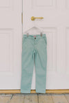 Sage Green Elio Chinos hang on a door, showcasing classic, effortless style with a slim fit and adjustable waist for fun, high-end comfort.