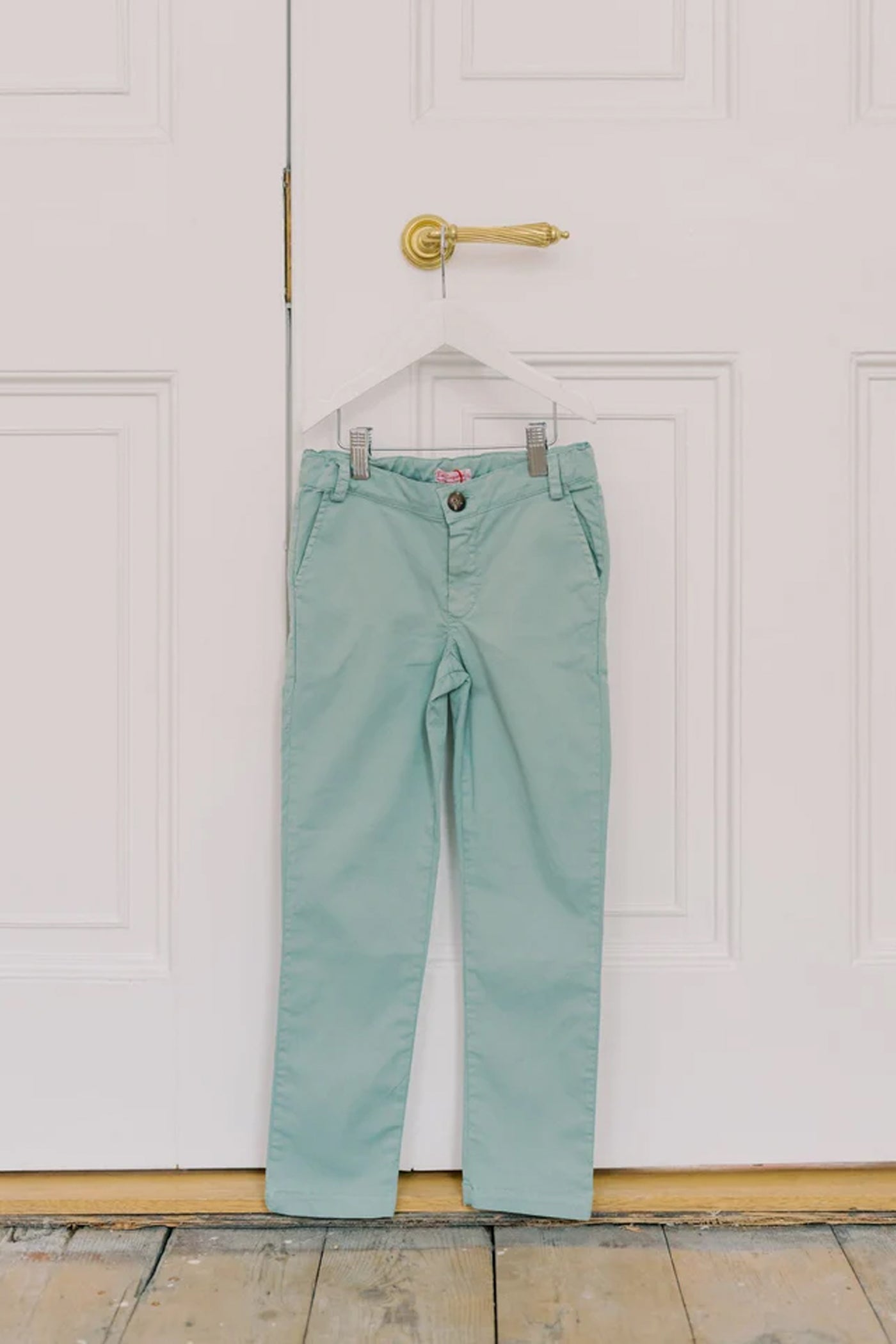 Sage Green Elio Chinos hang on a door, showcasing classic, effortless style with a slim fit and adjustable waist for fun, high-end comfort.