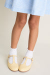 Child wearing blue-and-white gingham skirt with classic, high-end Pale Yellow Leather T-Bar Shoes, featuring effortless style and fun, ruffled white socks.