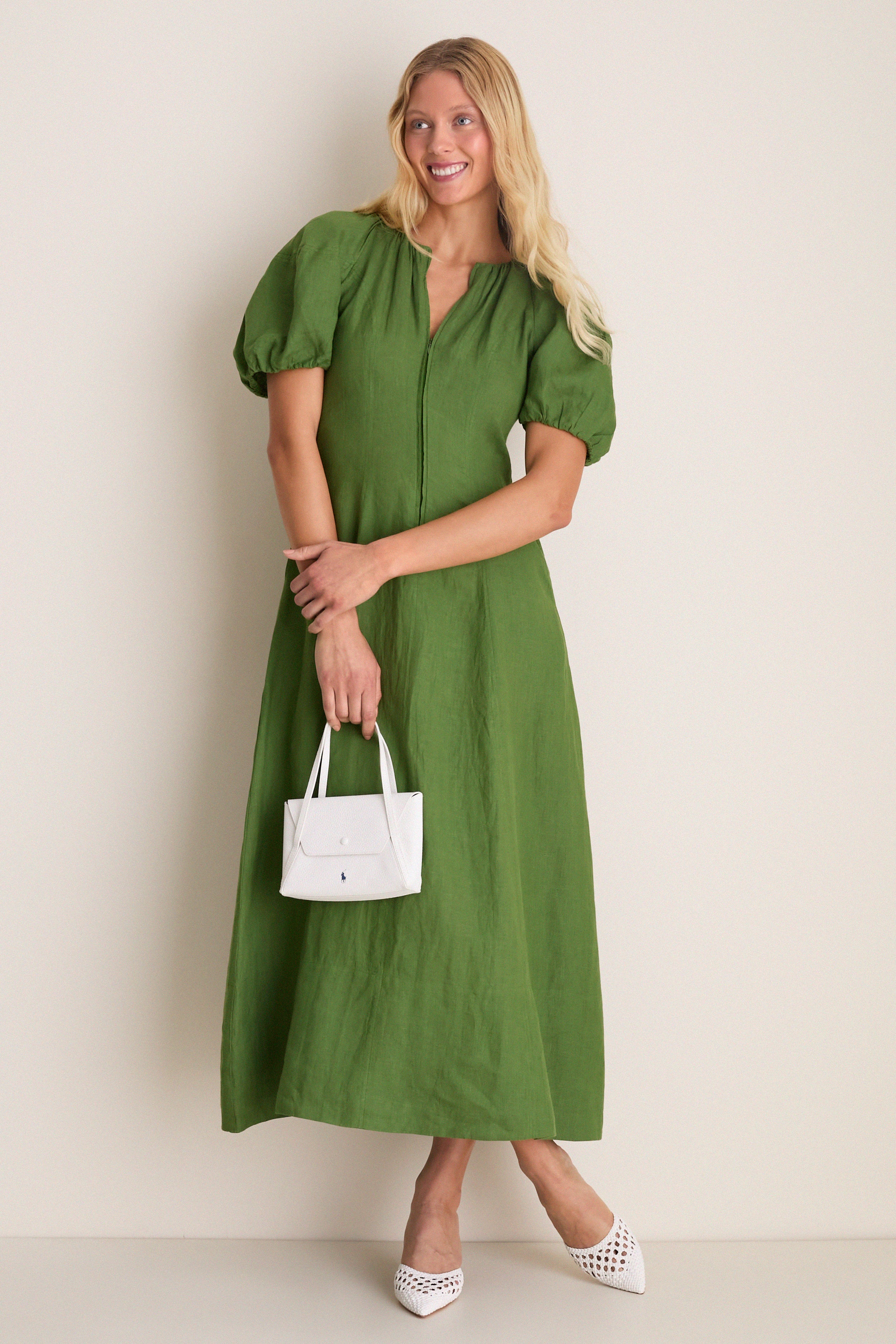 Woman in grass-green dress wearing classic White Tia Heels, featuring a woven silhouette and pointed toe, holding a white handbag. Effortless and high-end summer style.
