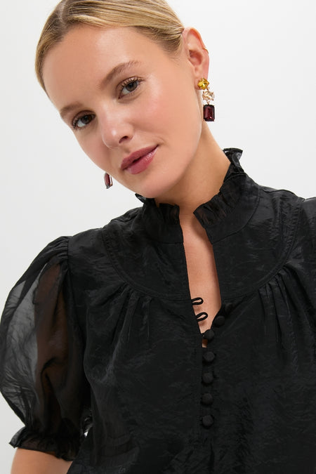 Black Chiffon Selena Blouse with sheer texture, ruffled collar, and covered buttons; worn by a model with elegant drop earrings, epitomizing classic, effortless high-end style.