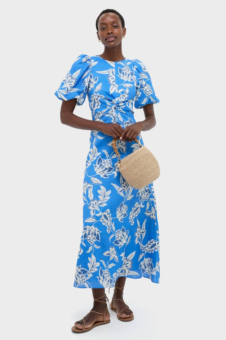 Woman in a Ren Cut Out Maxi Dress, featuring classic puff sleeves, a twist waist, and effortless floral design, paired with a fun straw handbag and high-end lace-up sandals.