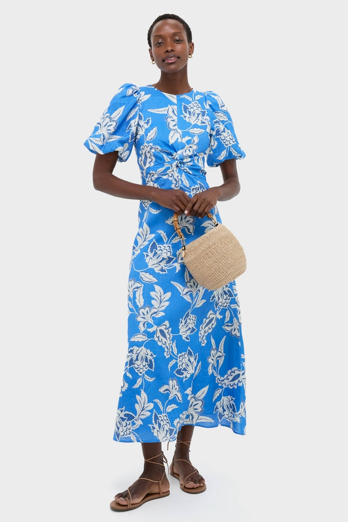 Woman in a Ren Cut Out Maxi Dress, featuring classic puff sleeves, a twist waist, and effortless floral design, paired with a fun straw handbag and high-end lace-up sandals.