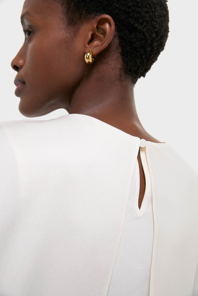 Cream Eliora Cape Top on a woman, showcasing its classic, asymmetric drape and effortless elegance with a keyhole button closure, paired with a fun, high-end gold hoop earring.