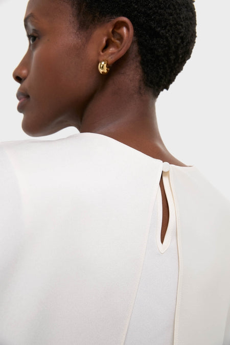 Cream Eliora Cape Top on a woman, showcasing its classic, asymmetric drape and effortless elegance with a keyhole button closure, paired with a fun, high-end gold hoop earring.