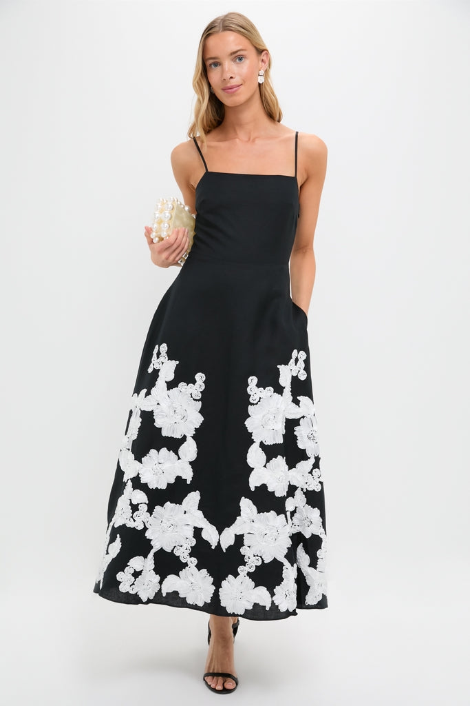 Model in a classic Black Multi Annabella Midi Dress with floral appliqués, holding a light-gold clutch. The dress exudes effortless, high-end style, perfect for fun occasions.