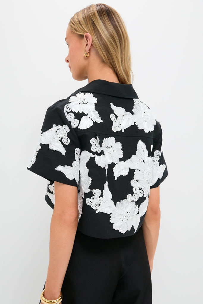 Woman in a classic Black Multi Callie Camp Shirt with white floral appliqués, embodying effortless elegance. She accessorizes with gold earrings and bracelets, exuding high-end, fun style.