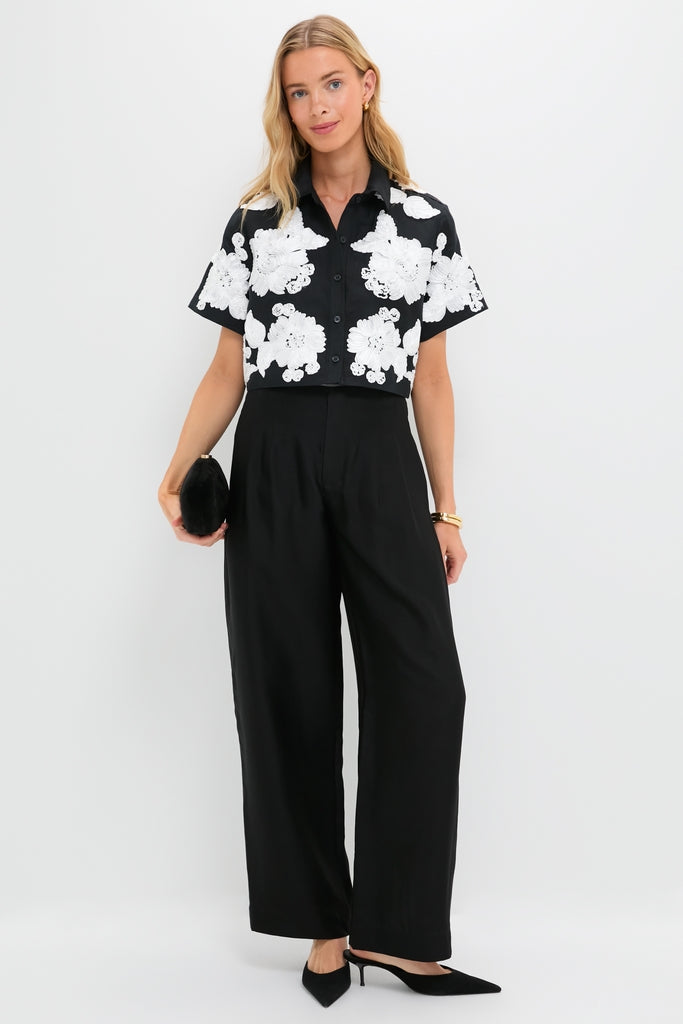 Woman models the classic Black Multi Callie Camp Shirt with floral appliqués, paired with black wide-leg pants and clutch, embodying an effortless, high-end style.