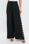 Model wearing Black Cayman Wide Leg Pants with high-waist, wide-leg design, featuring side slits with gold-tone buttons. Paired with a white top and strappy sandals for an effortless, high-end look.