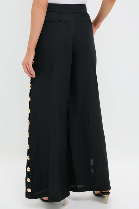 Woman in Black Cayman Wide Leg Pants with gold buttons, paired with a white top and black heeled sandals, embodying classic and effortless high-end style.