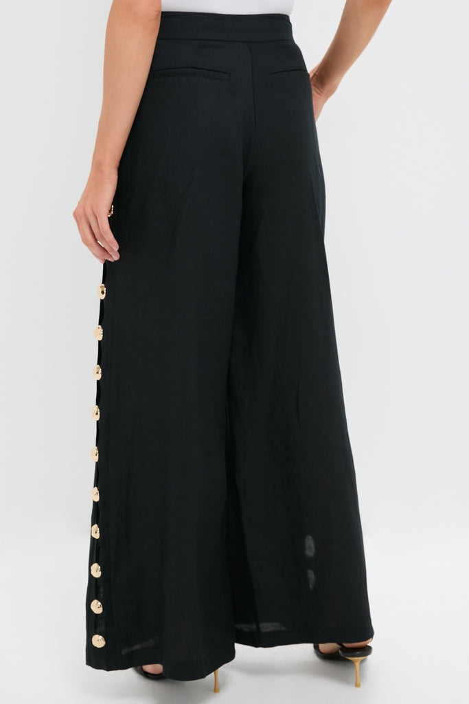 Woman in Black Cayman Wide Leg Pants with gold buttons, paired with a white top and black heeled sandals, embodying classic and effortless high-end style.