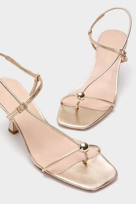 Champagne Metallic Triana Heels: Classic, high-end sandals featuring a thin ankle strap, T-strap, and decorative gold ball, blending effortless style with fun sophistication.