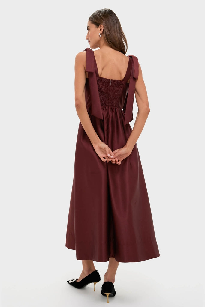 Woman in Oxblood Dupioni Bow Esme Dress, showcasing classic elegance with tied shoulder bows, a smocked back, and an A-line fit, paired with black heels.