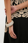 Person's lower torso wearing a black garment with swirling embroidery, showcasing the Sandy Pearl Rock Medium Bangle—an effortless, fun, high-end accessory in mottled gray and off-white tones.