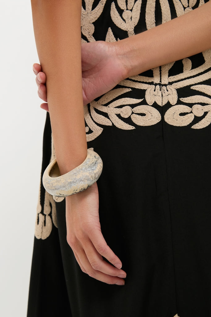 Person's lower torso wearing a black garment with swirling embroidery, showcasing the Sandy Pearl Rock Medium Bangle—an effortless, fun, high-end accessory in mottled gray and off-white tones.