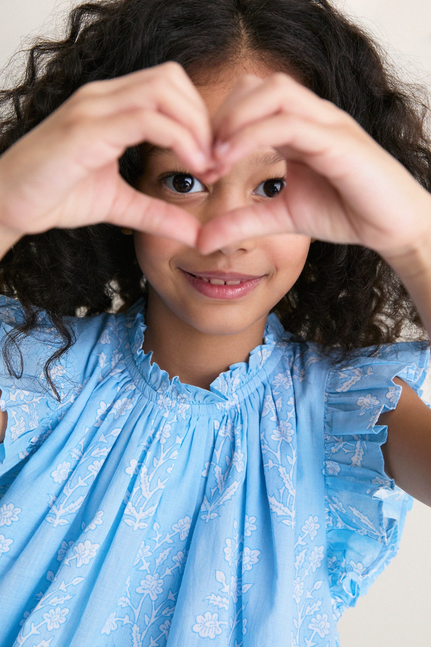 Young girl in a classic, light-blue Blue Dewdrop Blockprint Harper Dress with white flowers, creating a heart shape with her hands, embodying effortless fun and high-end style.