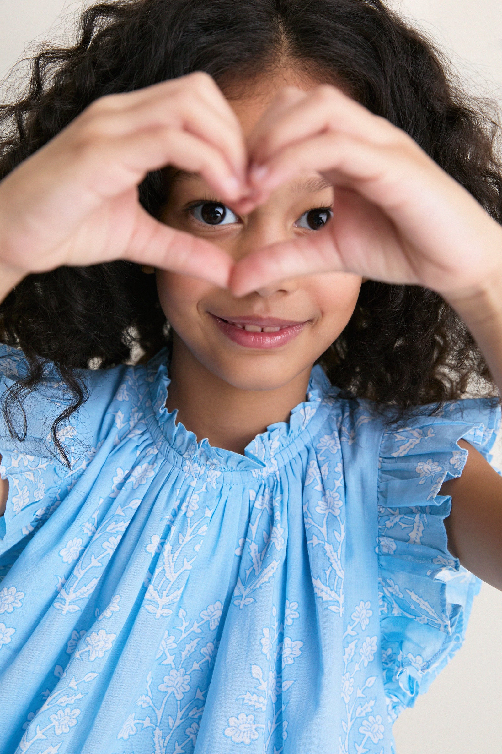 Young girl in a classic, light-blue Blue Dewdrop Blockprint Harper Dress with white flowers, creating a heart shape with her hands, embodying effortless fun and high-end style.