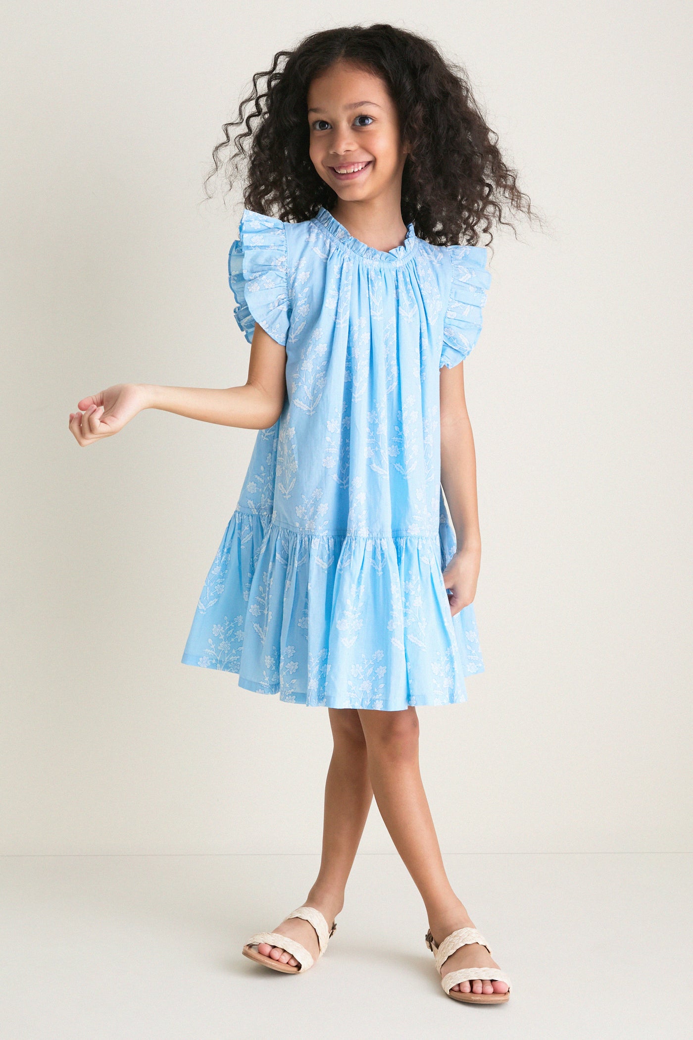 Young girl in a classic, effortless Blue Dewdrop Block Print Harper Dress with ruffle sleeves, posing confidently with braided-strap sandals for a fun, high-end look.
