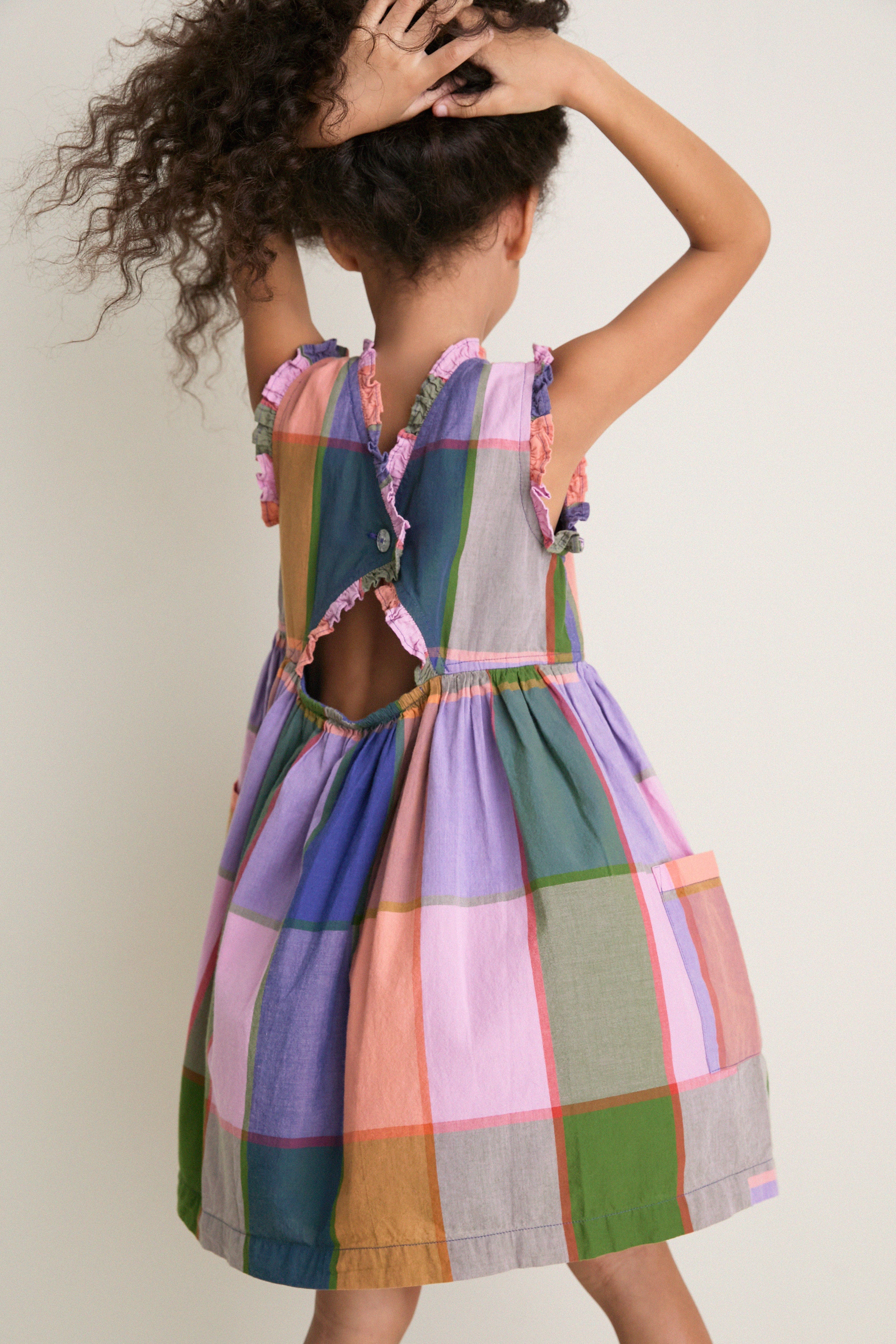 Young child in a classic, sleeveless Plaid Madras Alyssa Dress with ruffle trims, keyhole back, and raised arms, embodying effortless, fun style.
