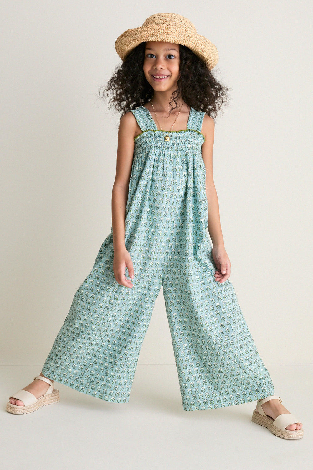 Young girl in a classic, effortless Blue Prairie Block Print Eleven Jumper, featuring a smocked bodice and wide-leg design, paired with a floppy-brim hat and espadrille sandals.