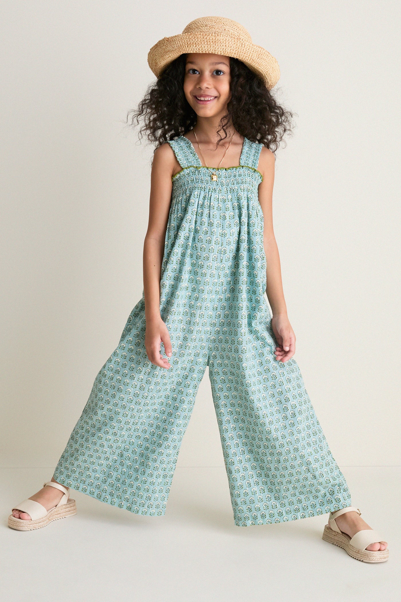 Young girl in a classic, effortless Blue Prairie Block Print Eleven Jumper, featuring a smocked bodice and wide-leg design, paired with a floppy-brim hat and espadrille sandals.